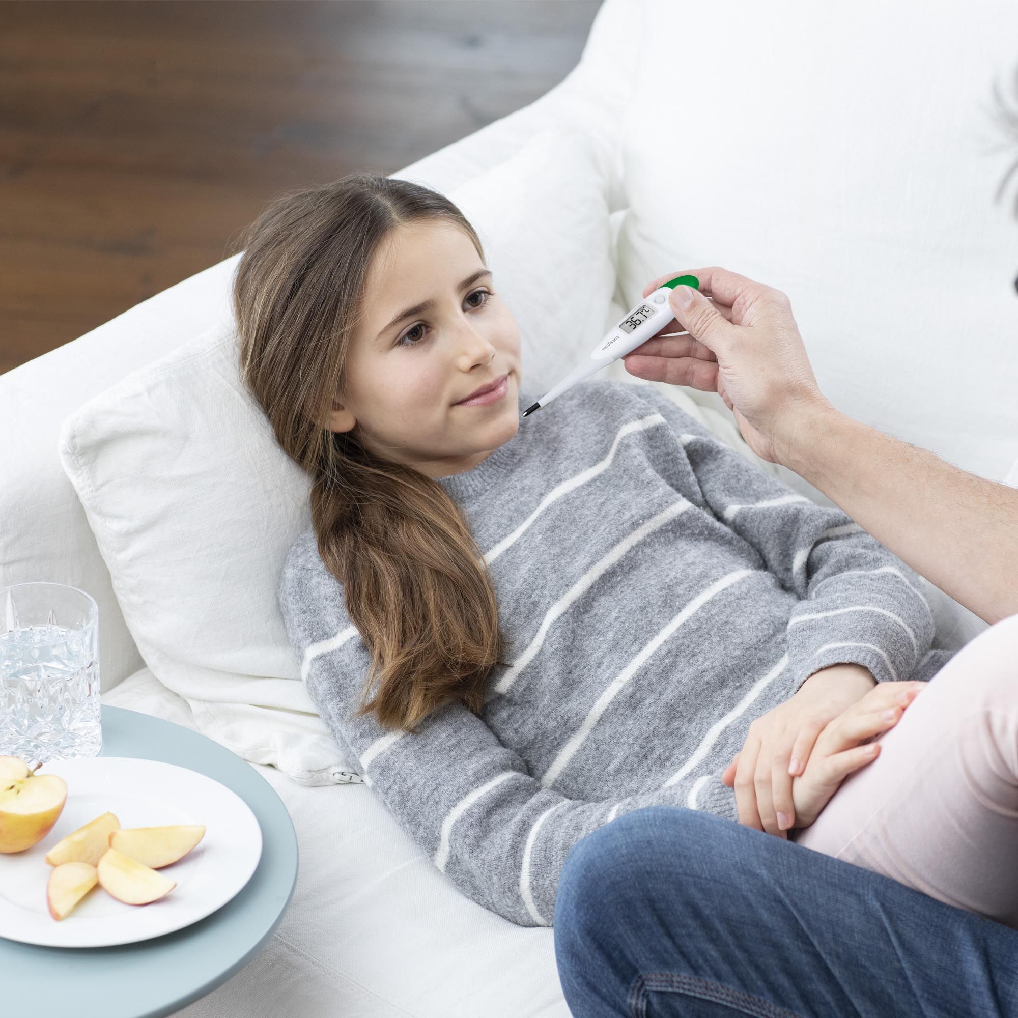 Kind liegt auf Sofa, Person misst Fieber mit digitalem Thermometer.