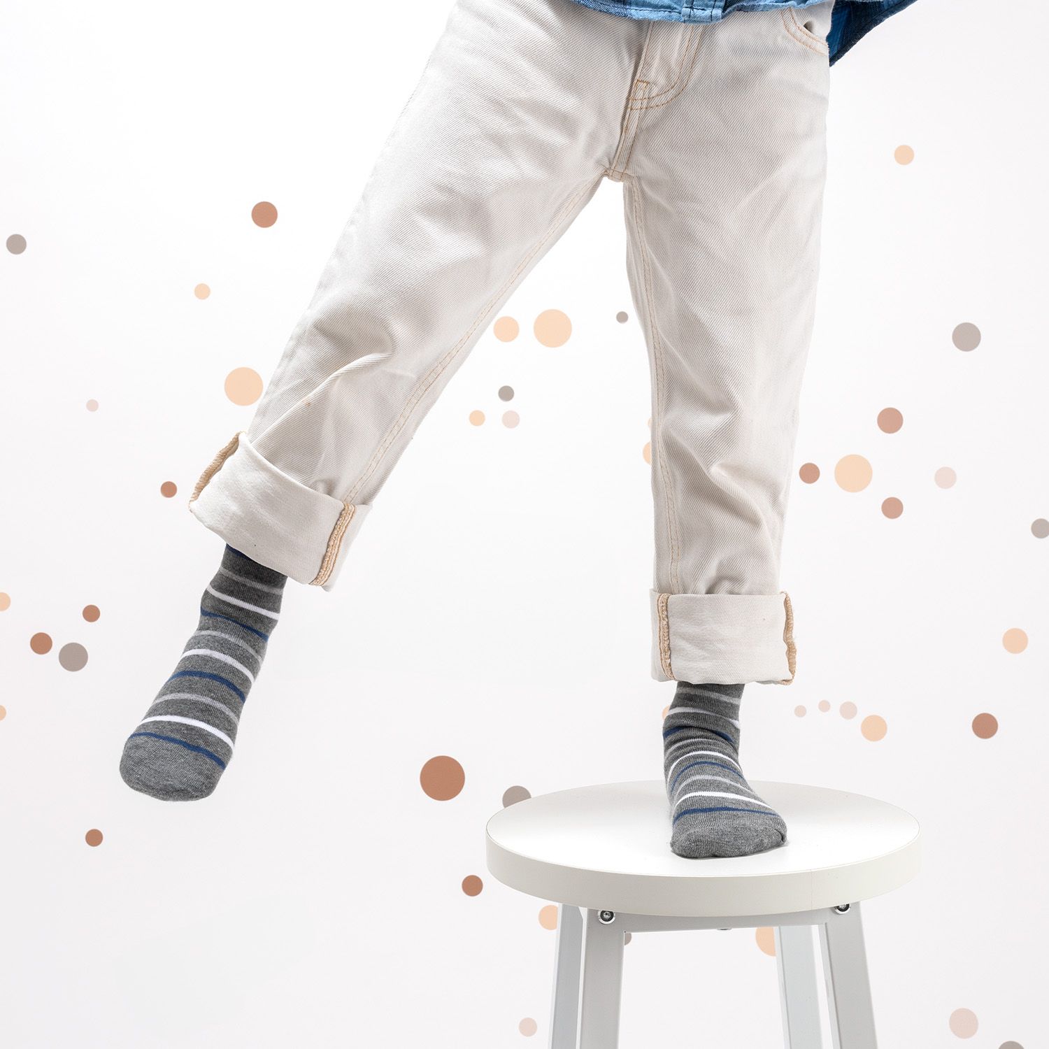 Kind steht auf einem Hocker, trägt gestreifte Socken in Grau und Blau. Weiße Hose.