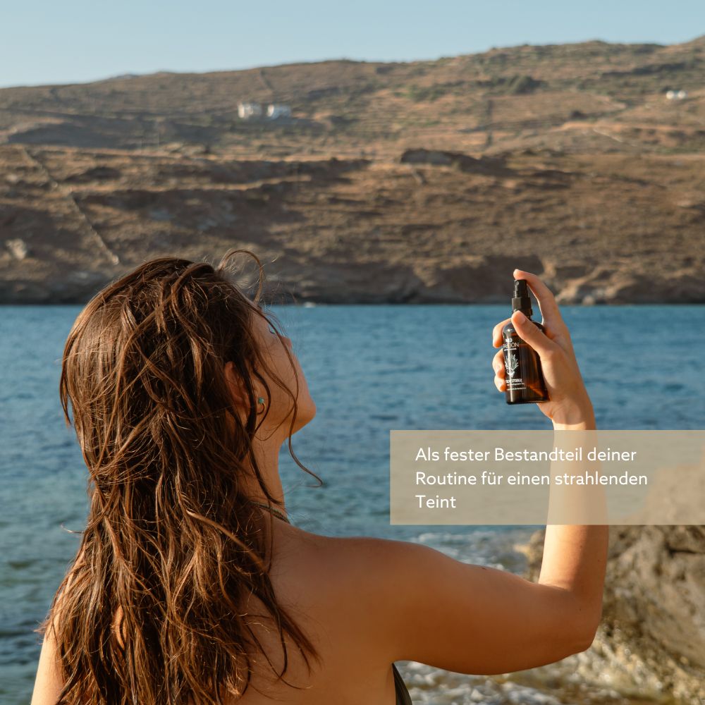 Frau am Meer hält eine braune Sprühflasche. Text: Als fester Bestandteil deiner Routine für einen strahlenden Teint.